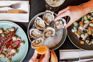 Flat lay of caucasian hands holding oysters with other seafood dishes on a dark table.